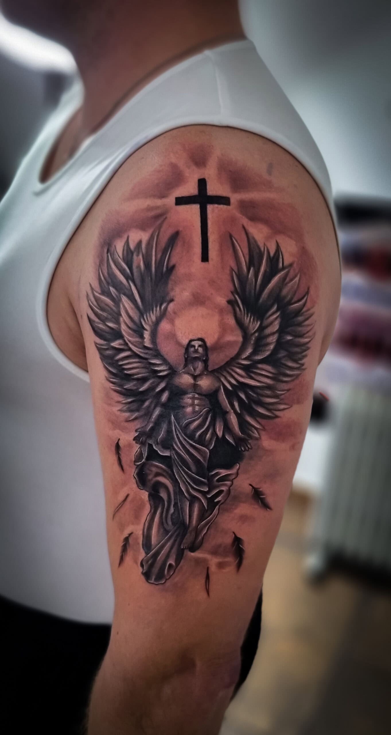Person mit weißem Tanktop und Engel-Tattoo am Oberarm, schwarzes Kreuz darüber; ausdrucksstark.