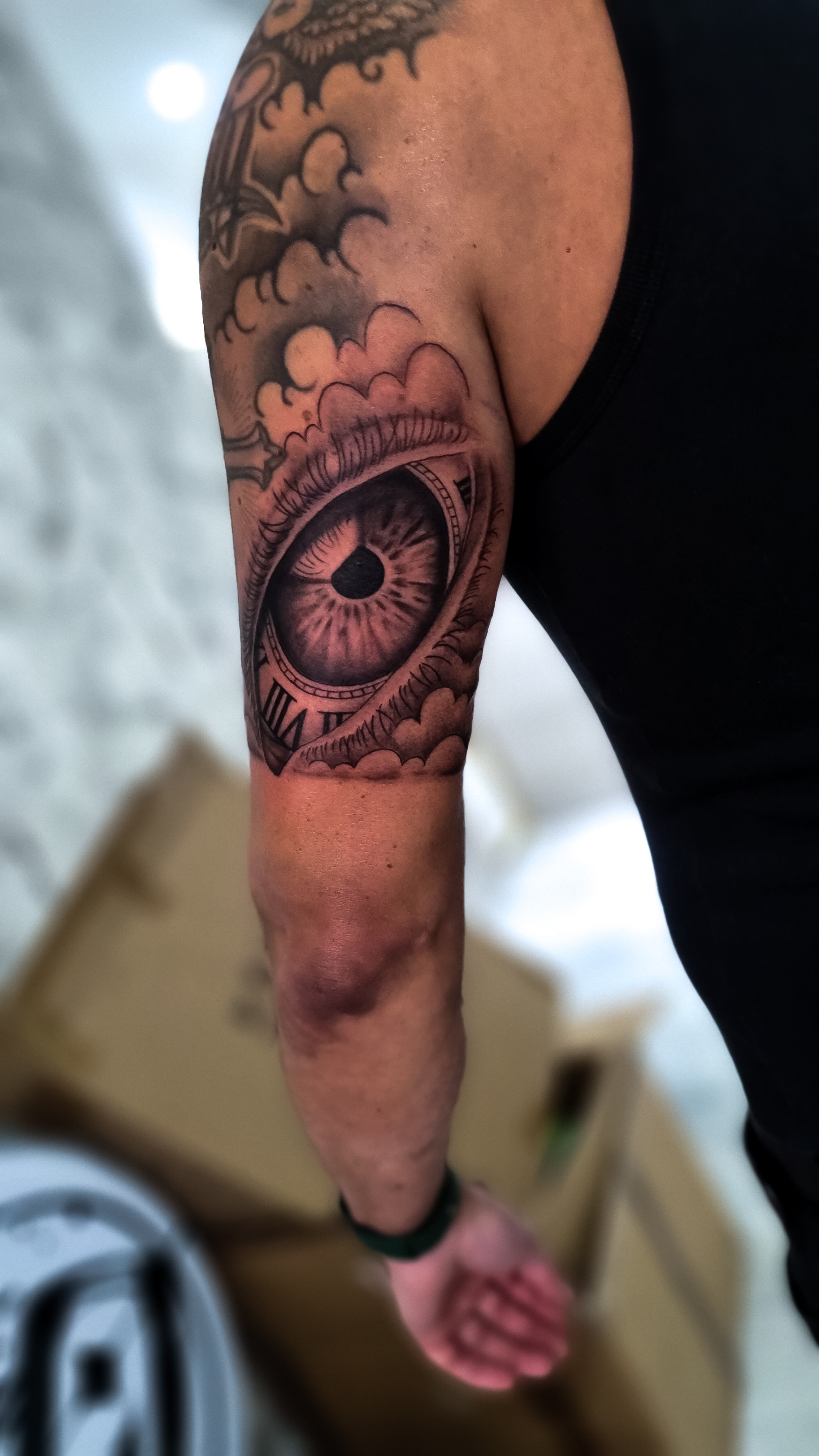 Ein männlicher Arm mit einem großem Augentattoo und Wolkendetails auf der Schulter.