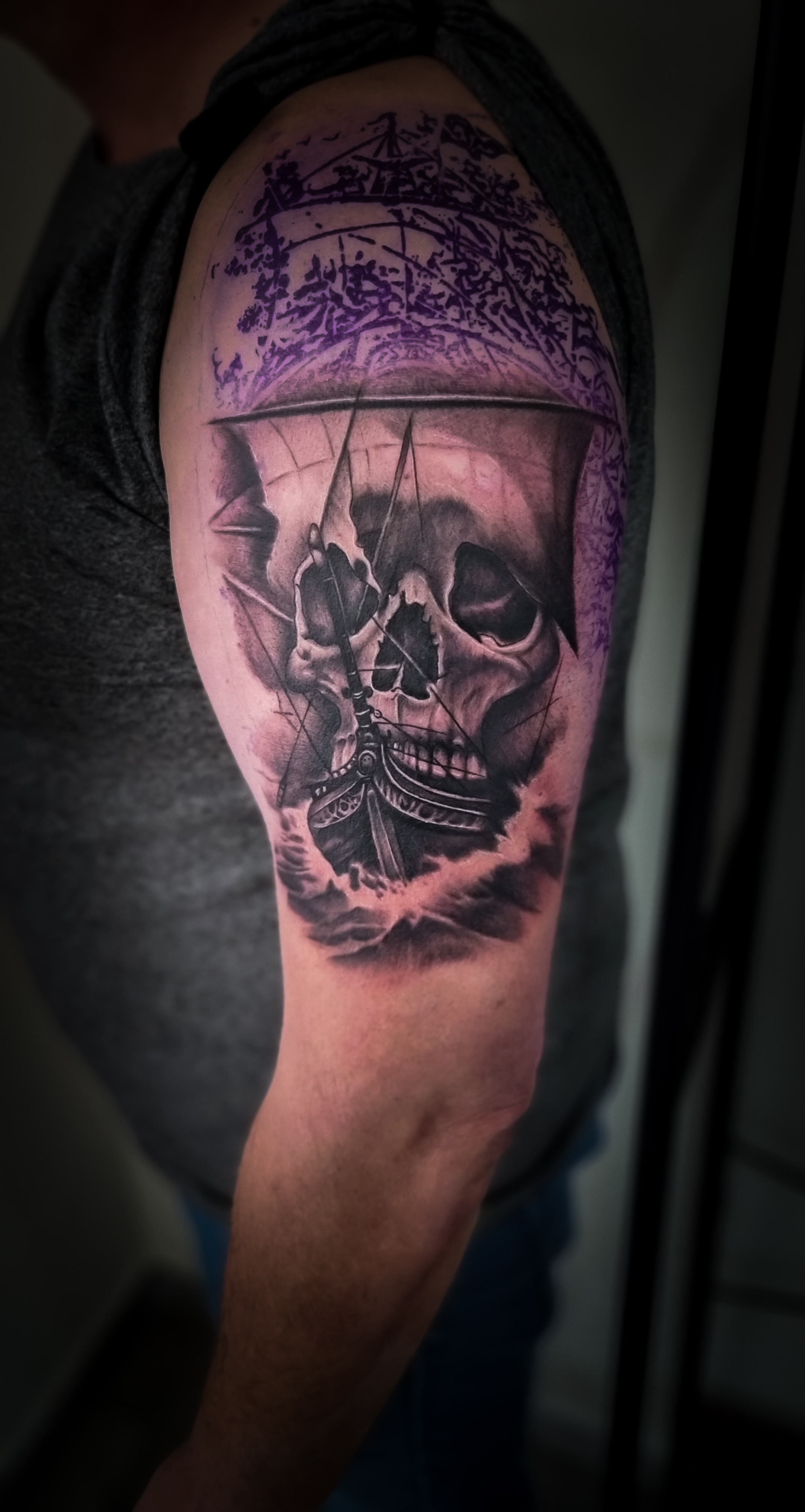 Oberarm-Tattoo mit Totenkopf und Schiff, auf männlichem Arm, graues T-Shirt, lebendige Farben.